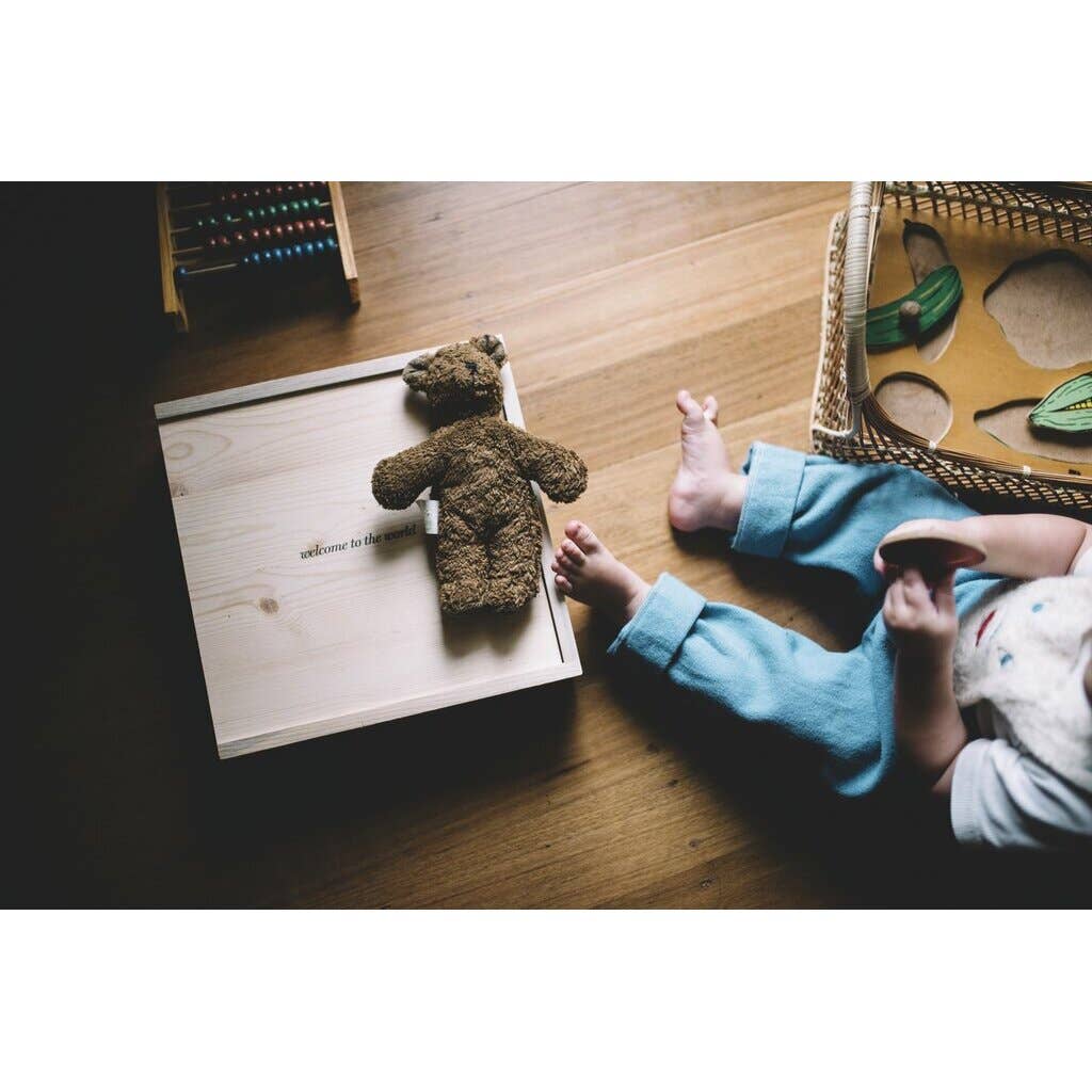A small child sits on a wooden floor with toys nearby. A GOTS-certified Senger Naturwelt Stuffed Animal - Brown Bear rests on a light wooden box beside the child, who wears eco-friendly quality blue pants.
