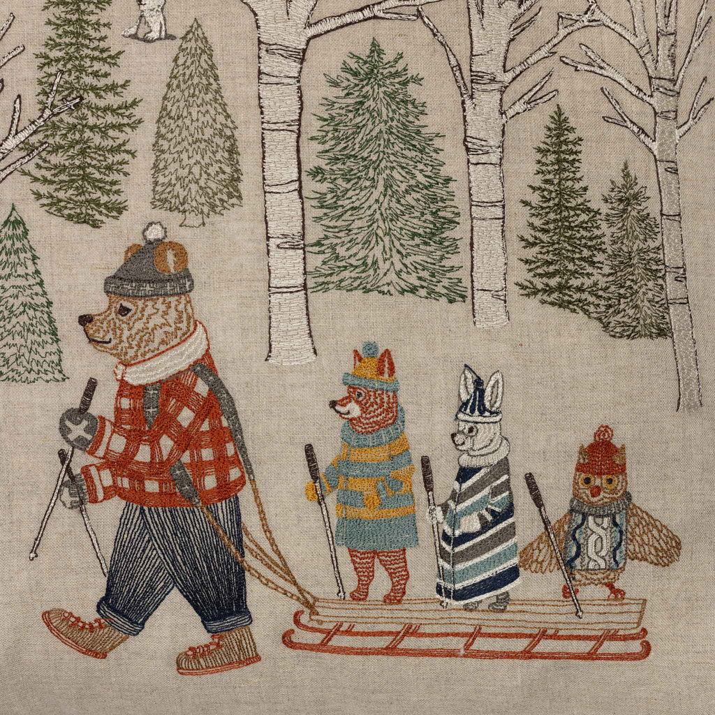 The Coral & Tusk Winter Fun Pillow features embroidered woodland animals—a bear towing a sled with a fox, rabbit, and owl—all in winter clothing with ski poles, set among snowy birch and evergreens for whimsical holiday decor.