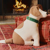 The Dog Cushion - Cooper, a plush handcrafted decor piece with brown and white patches and a green collar, sits on a red patterned rug near a red cushion, with a lit fireplace in the background.