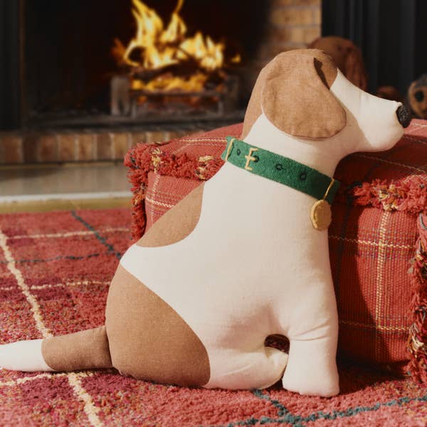 The Dog Cushion - Cooper, a plush handcrafted decor piece with brown and white patches and a green collar, sits on a red patterned rug near a red cushion, with a lit fireplace in the background.