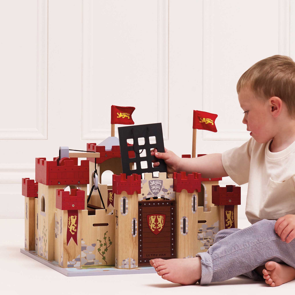A young child sits on the floor, engaged in imaginative play with the Lionheart Wooden Castle—a sustainable wooden toy adorned with red towers, flags, and a lion crest—while holding a black castle gate piece.