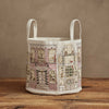 The Home for the Holidays Bucket, a canvas storage basket with two handles and embroidered buildings decorated with Christmas wreaths, garlands, and lights, rests on a wooden surface against a beige wall.