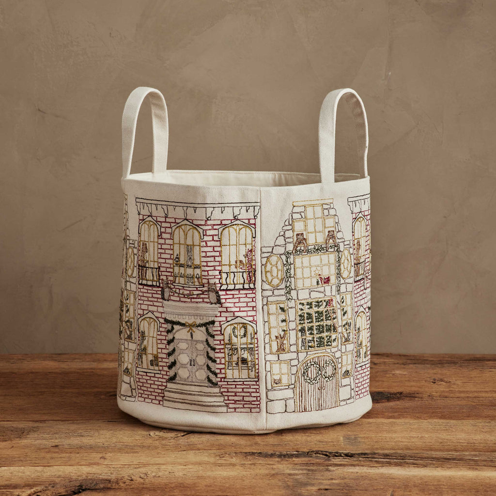 The Home for the Holidays Bucket, a canvas storage basket with two handles and embroidered buildings decorated with Christmas wreaths, garlands, and lights, rests on a wooden surface against a beige wall.