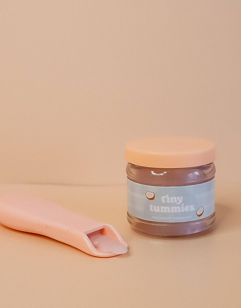 A jar labeled "Tiny Tummies - Coconut Yogurt" from Tiny Harlow sits on a beige surface next to a pink spoon, resembling a magic spoon, with yogurt on it. The jar features a light pink lid and a blue label decorated with cartoon coconut designs.