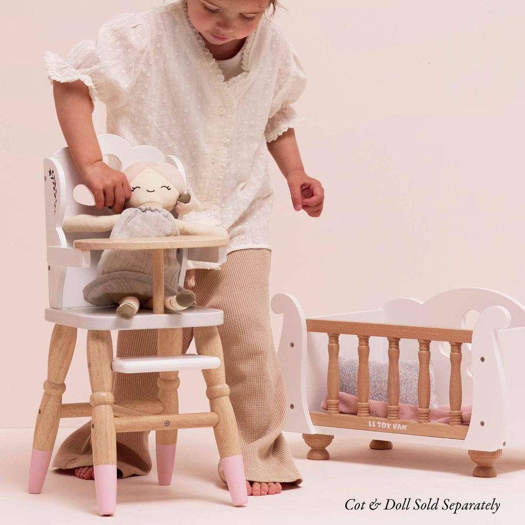 A child in light-colored clothes places a toy in the Dolls Wooden High Chair next to a matching crib. The pastel, eco-friendly toys add charm. Text in the corner reads: "Cot & Doll Sold Separately.