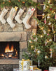 A cozy living room decorated for Christmas features a fireplace, a lit Christmas tree adorned with Yeti with Candy Cane Ornaments, and four stockings hanging on a stone wall. Wrapped gifts rest under the tree, while a warm fire glows invitingly in the hearth.