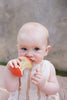 A blue-eyed baby in a sleeveless striped outfit bites into a red and yellow apple, teething just like they would with Pepa the Apple Teething Toy, while sitting against a soft, blurred background.