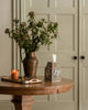 A round wooden table displays a vase with leafy branches, a lit orange candle, scissors, the Coral & Tusk Birdhouse Tissue Box Cover, and a tray. Beige double doors are in the background.