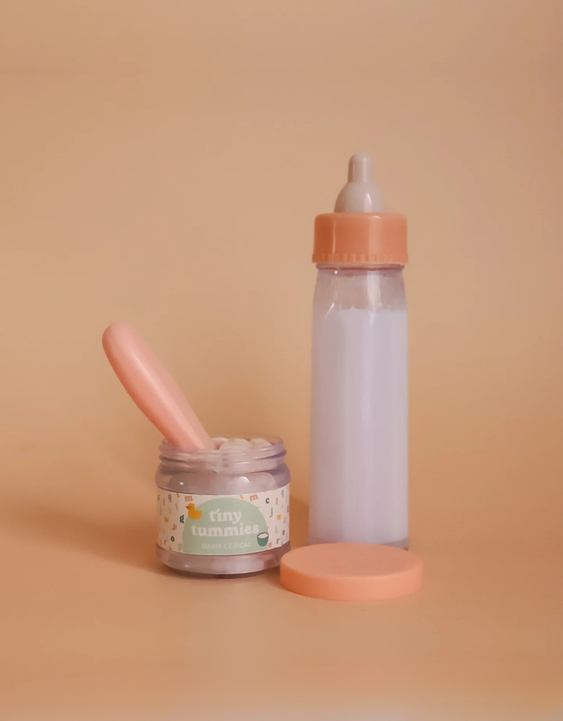 A jar from the My First Tiny Tummies Gift Set – Milk and Cereal, featuring a pink lid and spoon, is placed beside a magic milk bottle filled with light purple liquid on a beige background. The label on the jar reads "tiny tummies.