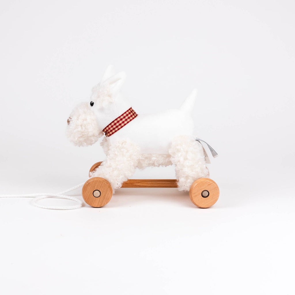 Pull-Along Lola is a white plush dog toy with a red checkered collar, mounted on four wooden wheels and a pull string—perfect for developmental play and an ideal toddler gift.