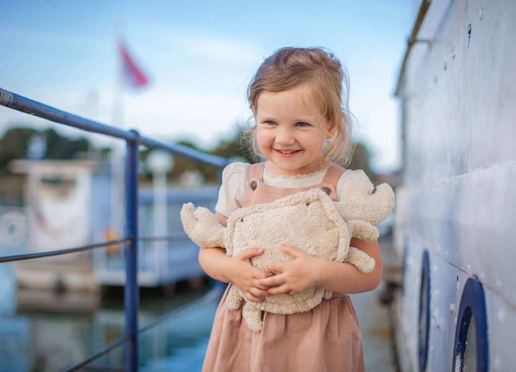 A young girl with light brown hair stands on a dock, smiling and hugging the Senger Naturwelt Cuddly Animal - Linen Crab, Small. She wears a light pink dress and white shirt. Boats and water are blurred in the background.