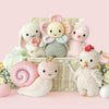 The Cuddle + Kind Baby Chicken, handmade in Peru for heirloom quality, is displayed with pastel animal friends and colorful eggs in a wicker basket adorned with flowers on a soft pink background.