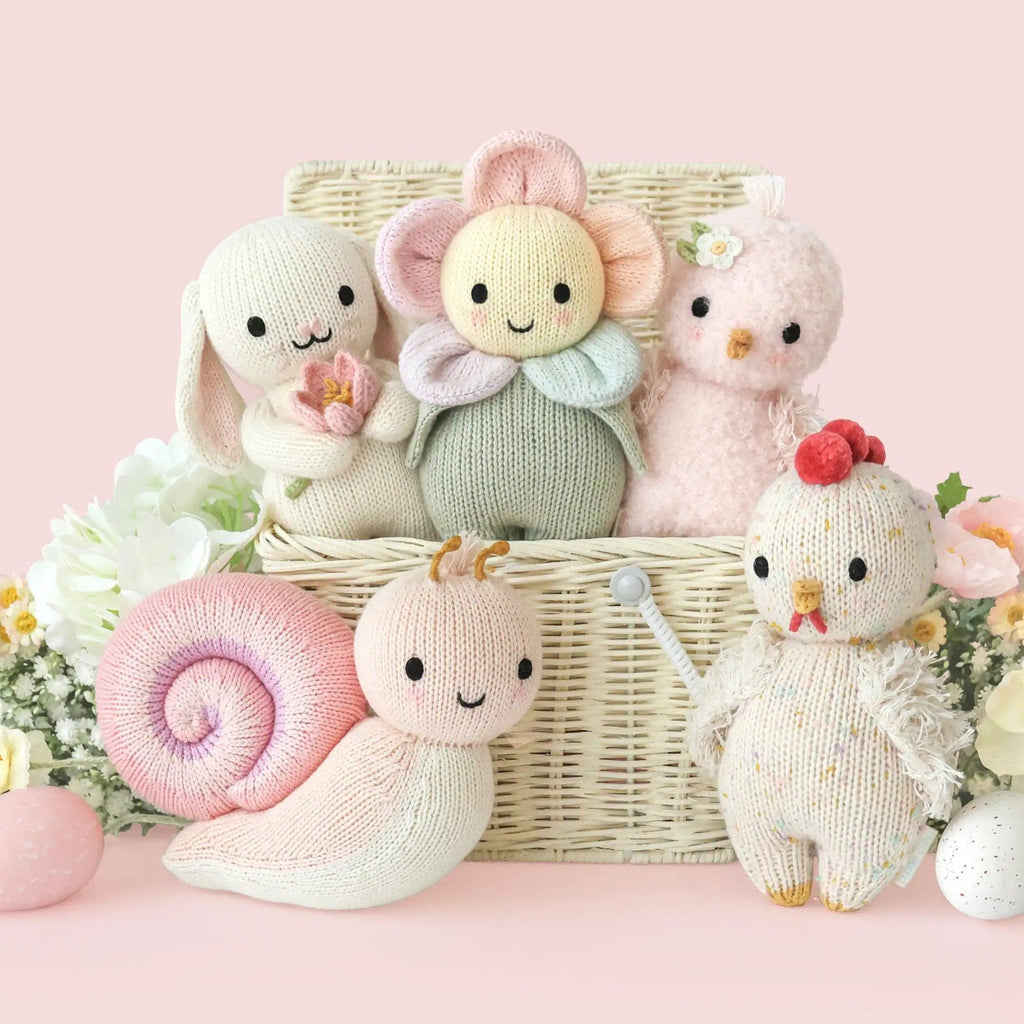 The Cuddle + Kind Baby Chicken, handmade in Peru for heirloom quality, is displayed with pastel animal friends and colorful eggs in a wicker basket adorned with flowers on a soft pink background.