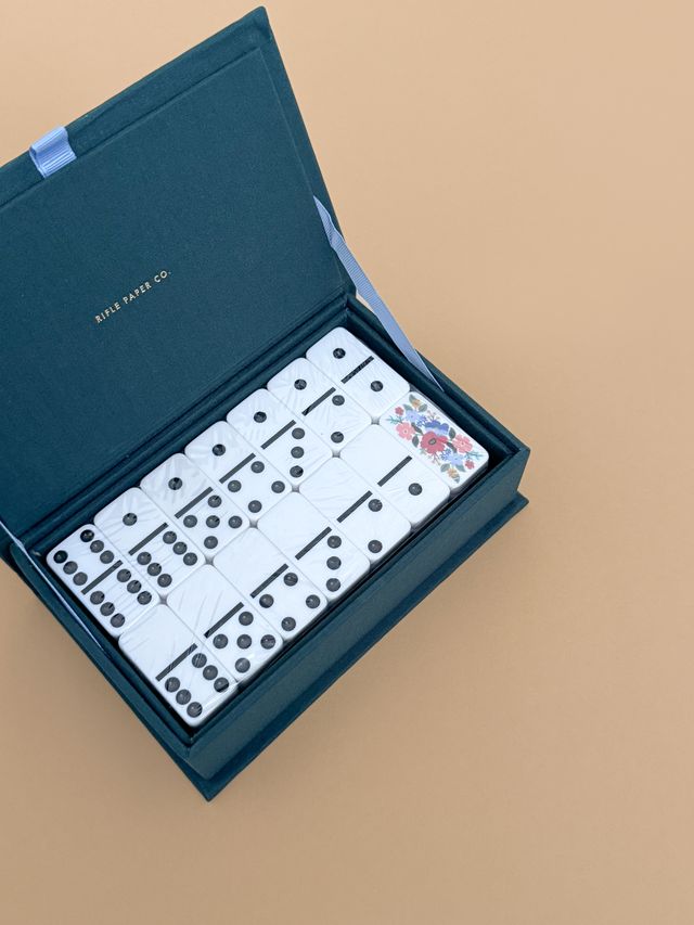 An open Strawberry Fields Dominoes Set box in dark teal displays white dominoes, one with a floral motif, all neatly arranged on a light tan surface.