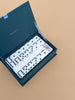 An open Strawberry Fields Dominoes Set box in dark teal displays white dominoes, one with a floral motif, all neatly arranged on a light tan surface.