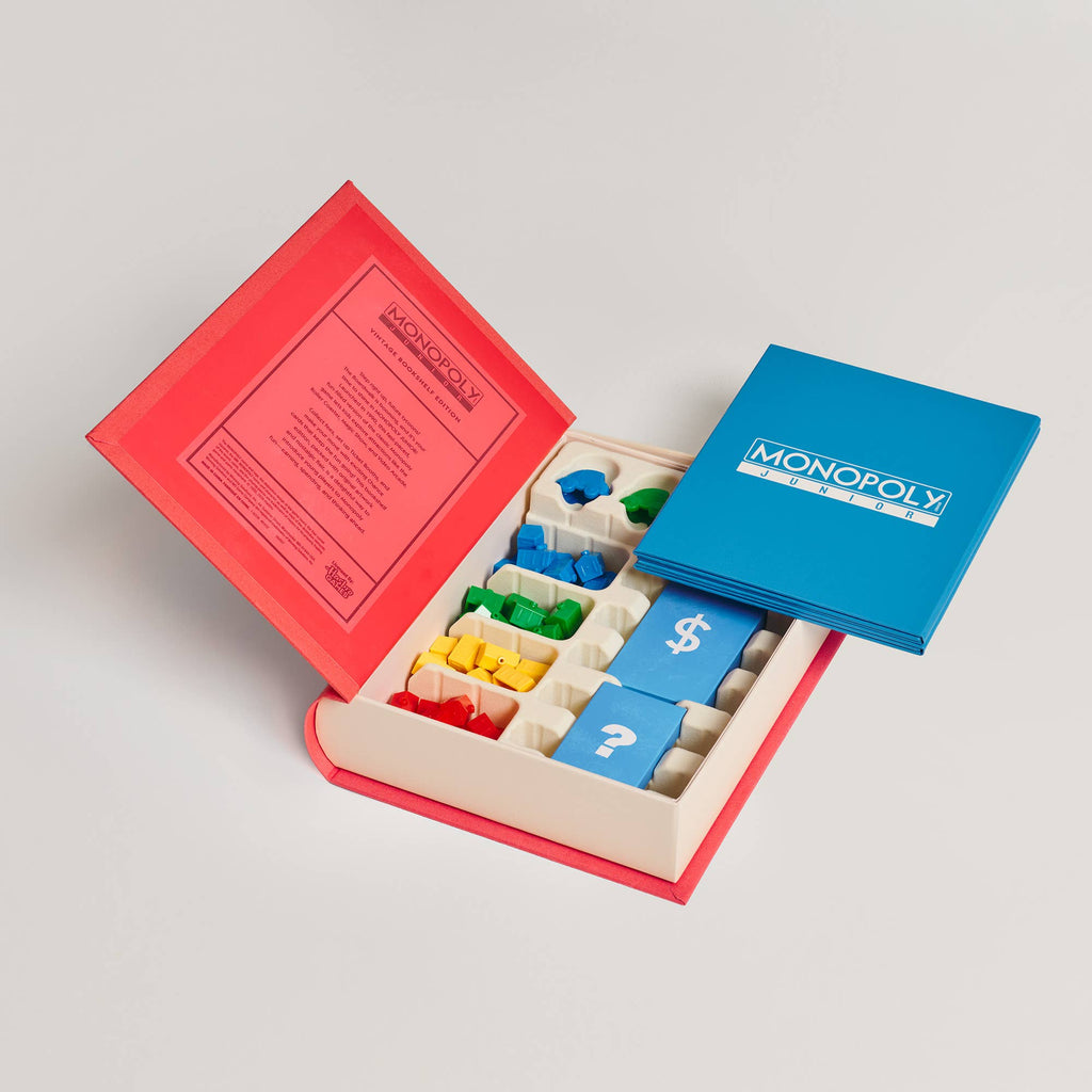 Open Monopoly Junior Vintage Bookshelf Edition box featuring a game board lid, plastic houses in blue, green, yellow, and red, and decks of cards with dollar sign and question mark icons.