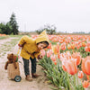 A young child in a yellow hooded coat bends to smell pink tulips in a field, with the Senger Naturwelt Stuffed Animal - Large Brown Bear and a basket on wheels beside them. Tulip fields extend beneath an overcast sky.