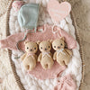 A pink knitted baby romper, mint bonnet, and a Cuddle + Kind Baby Honey Bear (Lavender Bow) are arranged on a cozy white blanket, with a heart-shaped sign reading "My First Valentine's Day" in the top right corner.