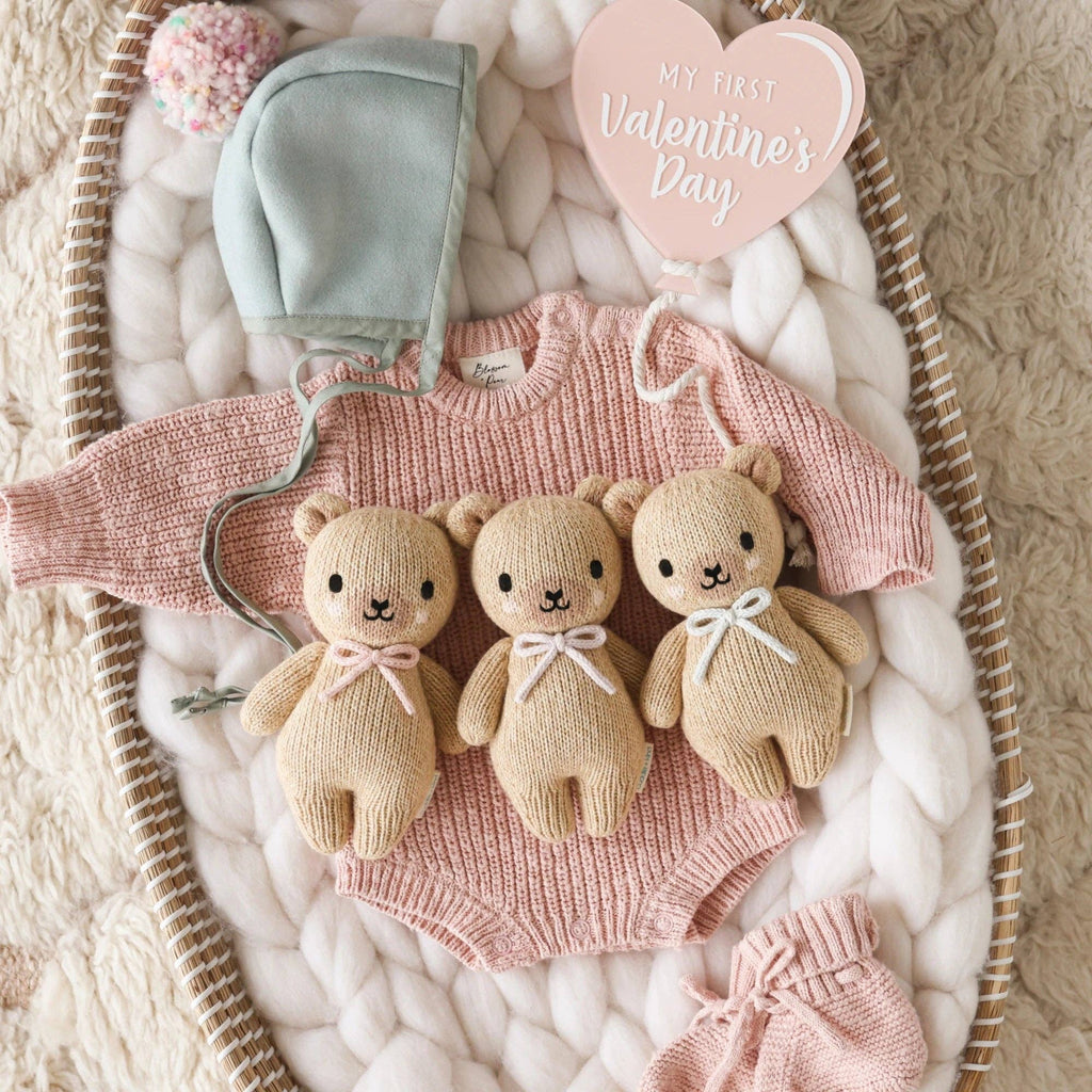 A pink knitted baby romper, mint bonnet, and a Cuddle + Kind Baby Honey Bear (Lavender Bow) are arranged on a cozy white blanket, with a heart-shaped sign reading "My First Valentine's Day" in the top right corner.