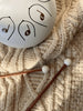 A Steel Tongue Drum with musical notes rests on a beige knitted sweater. Two wooden mallets with white tips lie nearby on the textured fabric, creating a delightful musical toy instrument for kids.