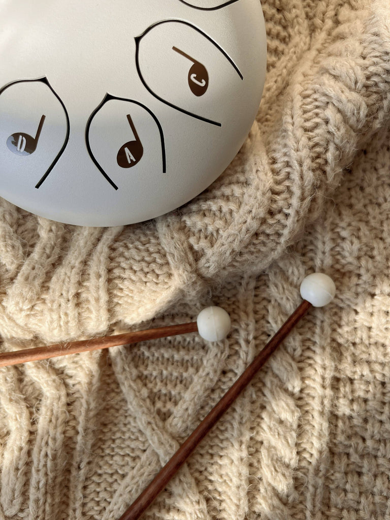 A Steel Tongue Drum with musical notes rests on a beige knitted sweater. Two wooden mallets with white tips lie nearby on the textured fabric, creating a delightful musical toy instrument for kids.