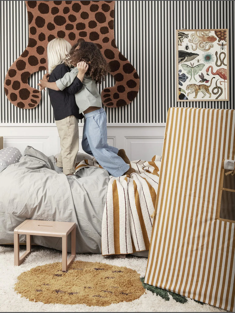 Two children playing in a lively room, one jumping into the other's arms for a hug near a bed adorned with a Ferm Living Fruiticana Tufted Pineapple Rug. The room has striped walls, a large bear wall decoration.