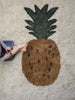 A child sits next to a large Ferm Living Fruiticana Tufted Pineapple Rug on a light carpeted floor, with only the lower part of their body visible. The rug features a textured brown base with black details and a green.