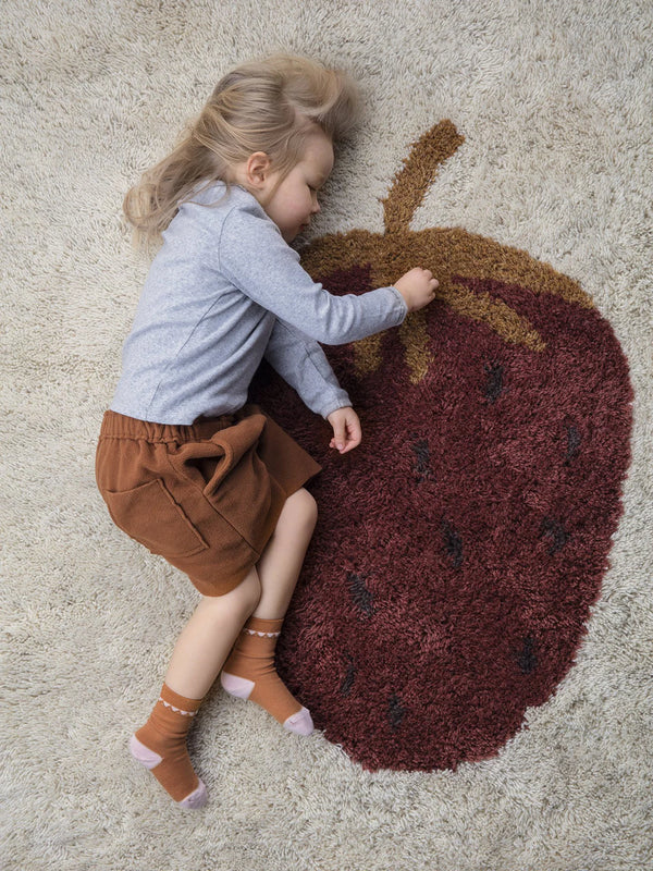 A young child with blonde hair peacefully napping on a large, fluffy Ferm Living Fruiticana Tufted Strawberry Rug, evoking a cozy and serene atmosphere.