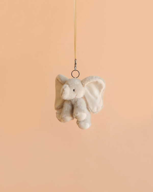 The Bukowski - August Elephant XL Bag Charm, a plush elephant keychain, hangs from a beige ribbon against a plain light peach background, making it an adorable accessory for any bag charm enthusiast.