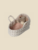 A soft toy resembling a small mouse is tucked into a cozy, patterned fabric basket. The mouse is covered with a pink, textured blanket and has a similarly patterned pillow, creating a snug and comforting scene akin to a Maileg Carry Cot, Baby.