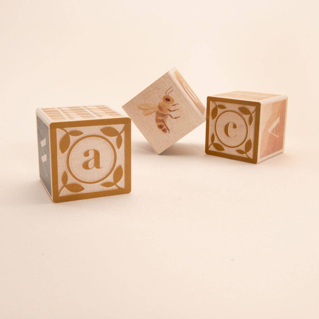 The Uncle Goose Watercolor ABC Blocks feature three sustainably crafted basswood cubes placed on a light background. One cube showcases the letter "a" adorned with decorative leaves, another presents an illustration of a bee, and the third highlights the letter "c" encircled by a paintbox design. These perfectly crafted learning tools are designed to inspire young minds.