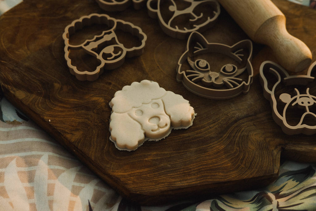 The Pet Friends Faces Cutter Set - Eco Playdough Tool includes dog, cat, and cow-shaped cutters, along with dog-shaped cookie dough and a rolling pin, all displayed on a wooden cutting board—perfect for sensory play and creative fun with playdough tools.
