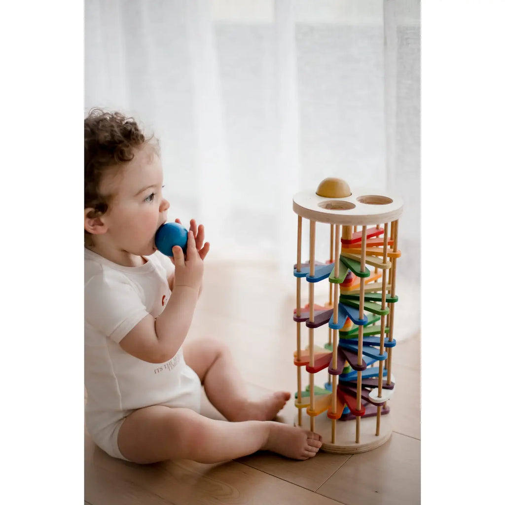 A toddler with curly hair sits on a wooden floor next to the Pound A Ball Tower, a rainbow track toy made from non-toxic materials. Dressed in a white onesie and holding a blue ball, the child brings it to their mouth. Soft natural light filters through sheer white curtains in the background, highlighting their developing motor skills.