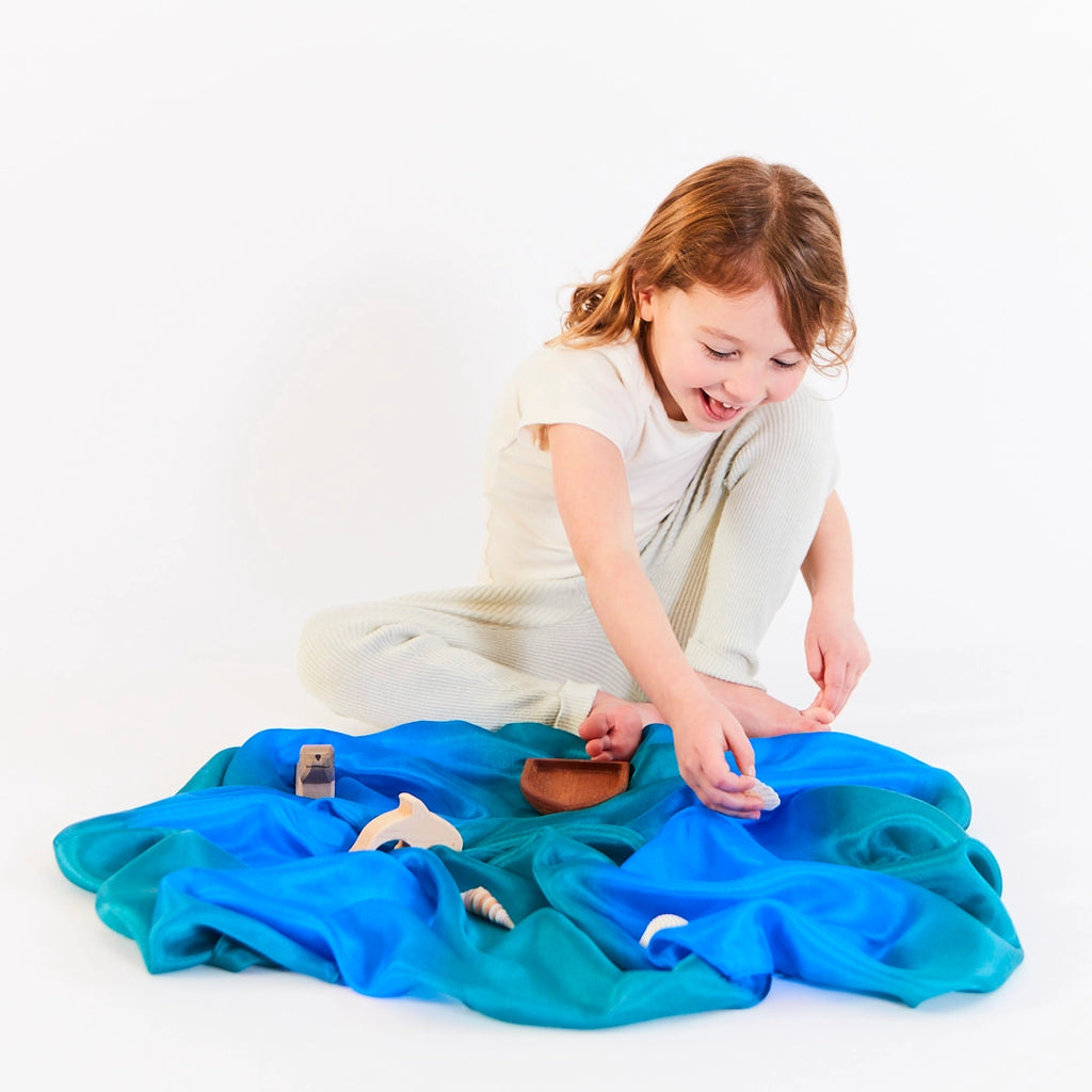 A young child sits on the floor, joyfully immersed in imaginative play, exploring various wooden animal figures spread across colorful Sarah's Silk Enchanted Playsilk - Ocean beneath them. The plain white background accentuates their delightful exploration.