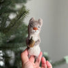 A hand holds the Felt Finger Puppet: Gray Squirrel, featuring gray fur, a brown nose, and white belly, holding a small brown object with a blurred pine branch in the background—an eco-friendly gift for kids.