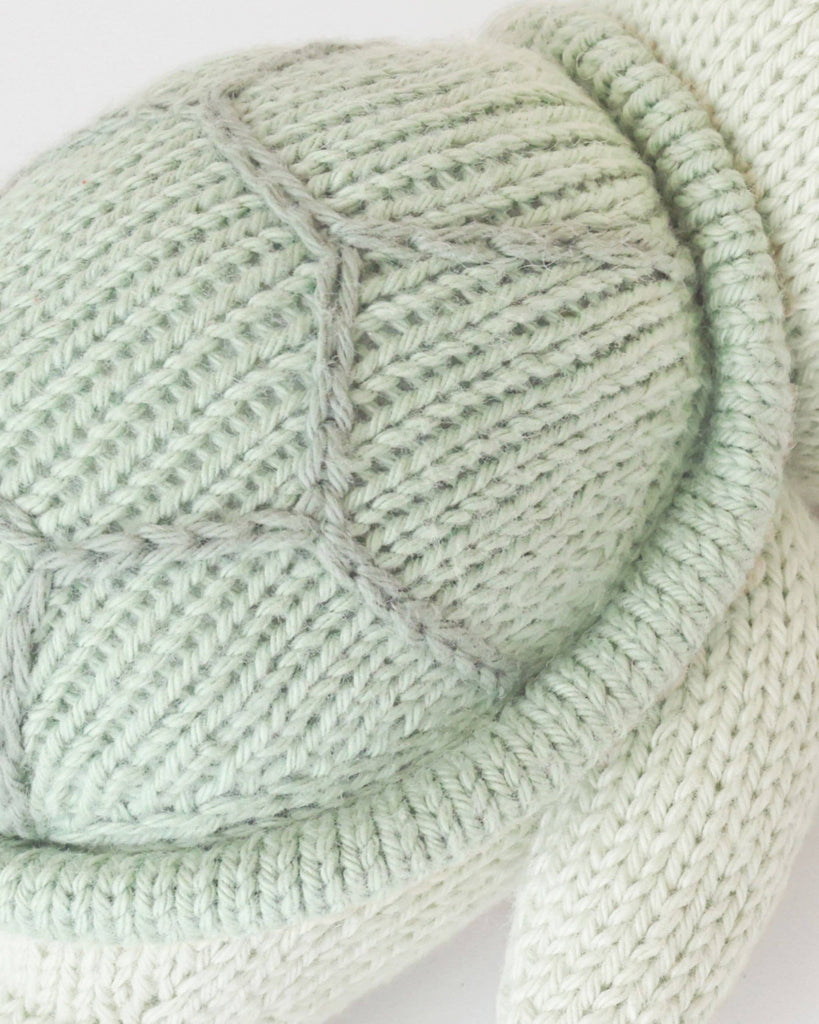 Close-up of the Cuddle + Kind Baby Sea Turtle (Kelp Green), a soft, hand-knit toy featuring a textured shell with gray stitching and part of its flipper—a charming, heirloom-quality piece.