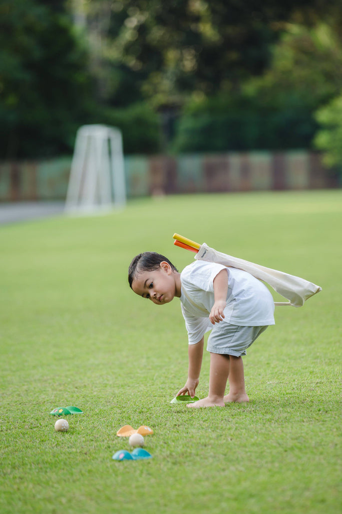 A young child wearing a white shirt and shorts picks up a green ball on the grass near sports gear and a goalpost, carrying their Mini Golf Set kids golf clubs over their shoulder.