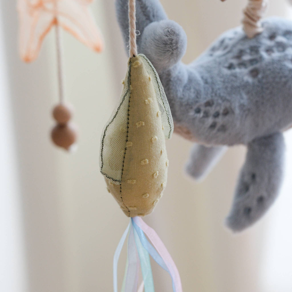 A close-up of the Nautical Dreams Mobile shows a soft, yellow, pear-shaped rattle toy with a dotted texture hanging by a string. A gray plush animal and a peach ornament appear in the background—perfect for nursery decor.