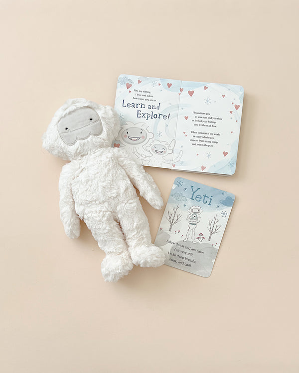 A Slumberkins Yeti Kin + Lesson Book - Mindfulness lies next to cards with illustrations and text about yetis on a light beige background, designed as stuffed animals to reduce anxiety.