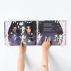 A child's hands holding open a Slumberkins "The Costume Comeback" Halloween Hardcover Book with colorful illustrations of children in various costumes dancing among stars and planets against a dark sky. Text appears on the left page.