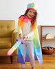 A smiling child, immersed in pretend play, wears a rainbow-colored cape and crown while brandishing Sarah's Silk Soft Sword indoors. They stand next to a yellow armchair and a shelf with toys in their cozy room adorned with rainbow silk accents.