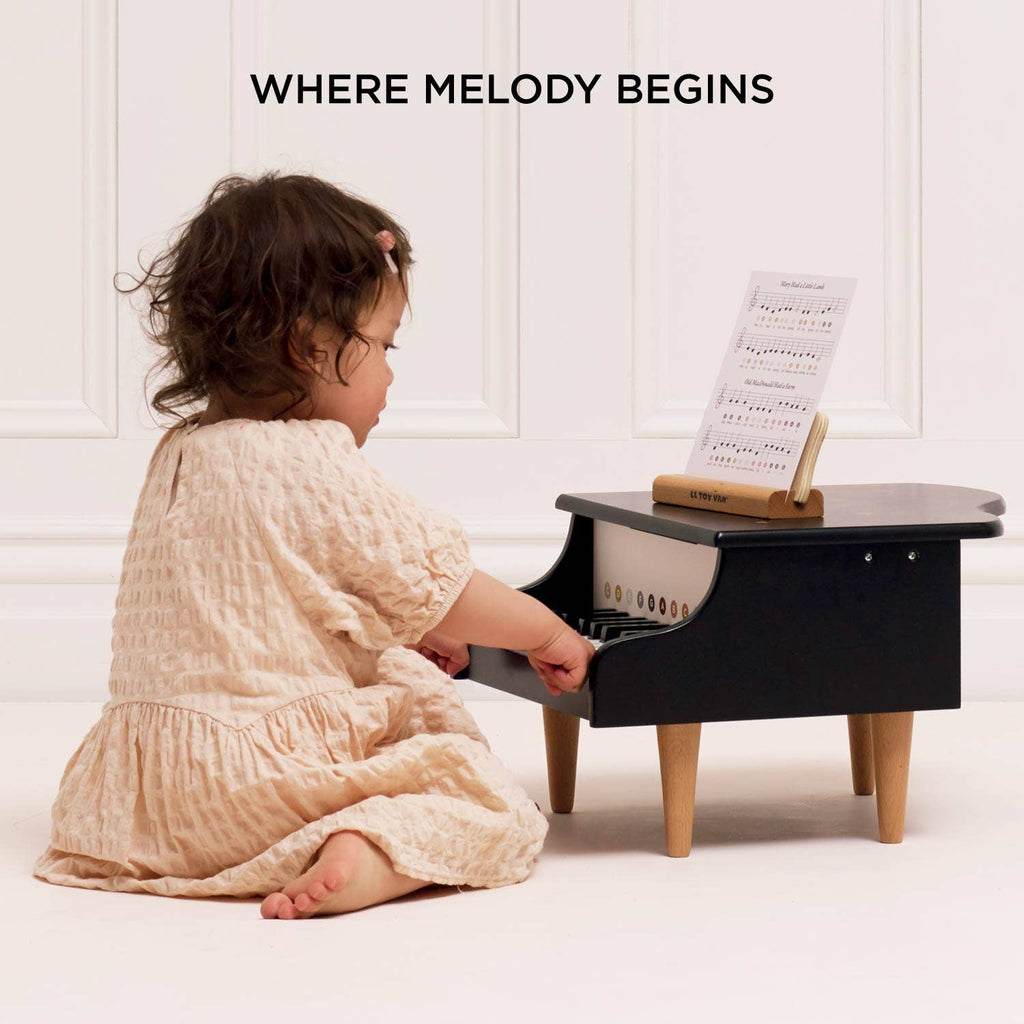 A young child in a light pink dress plays the Wooden Grand Piano Toy made from FSC-certified wood with sheet music, seated on the floor against a white wall. Text above says, "WHERE MELODY BEGINS.