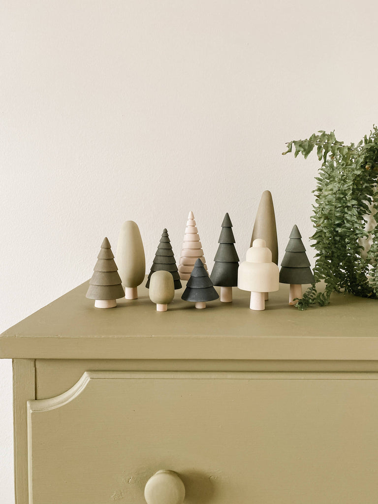 A charming collection of the Wooden Forest Set by SABO concept features small, stylized trees crafted from wood in diverse green hues and natural finishes, elegantly arranged on a light green dresser. To the right, a small plant with lush green leaves is partly visible, complementing the theme of imaginative play.