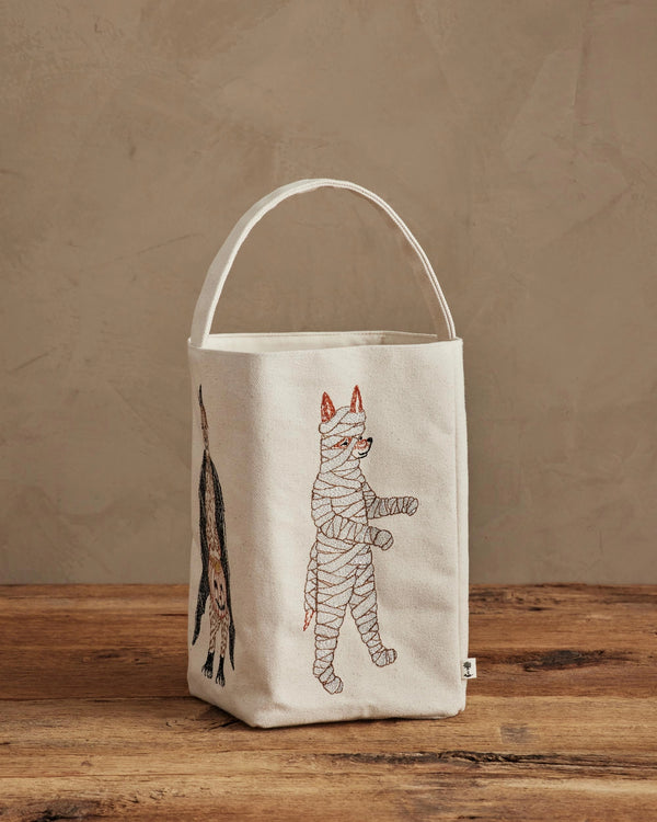 A cream-colored Coral & Tusk Halloween Trick Or Treat Bag (ships in one week) with embroidered costumed cats—a mummy and an upside-down black cat—rests on a wooden surface against a beige wall, perfect for Halloween.