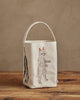 A cream-colored Coral & Tusk Halloween Trick Or Treat Bag (ships in one week) with embroidered costumed cats—a mummy and an upside-down black cat—rests on a wooden surface against a beige wall, perfect for Halloween.