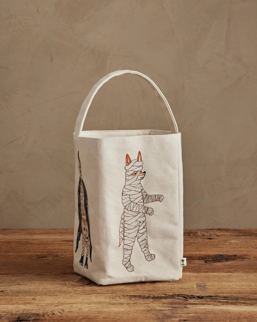 A cream-colored Coral & Tusk Halloween Trick Or Treat Bag (ships in one week) with embroidered costumed cats—a mummy and an upside-down black cat—rests on a wooden surface against a beige wall, perfect for Halloween.