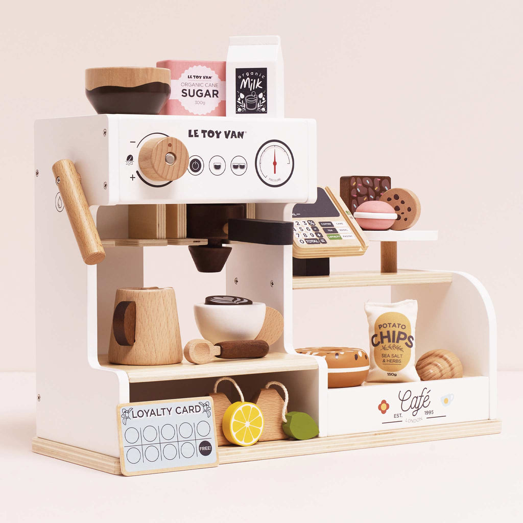 The Barista Café Shop features a wooden coffee machine, cups, milk, sugar, pastries, potato chips, a loyalty card, lemons, and a cash register on white shelves—perfect for endless pretend café play. Ships in one week.