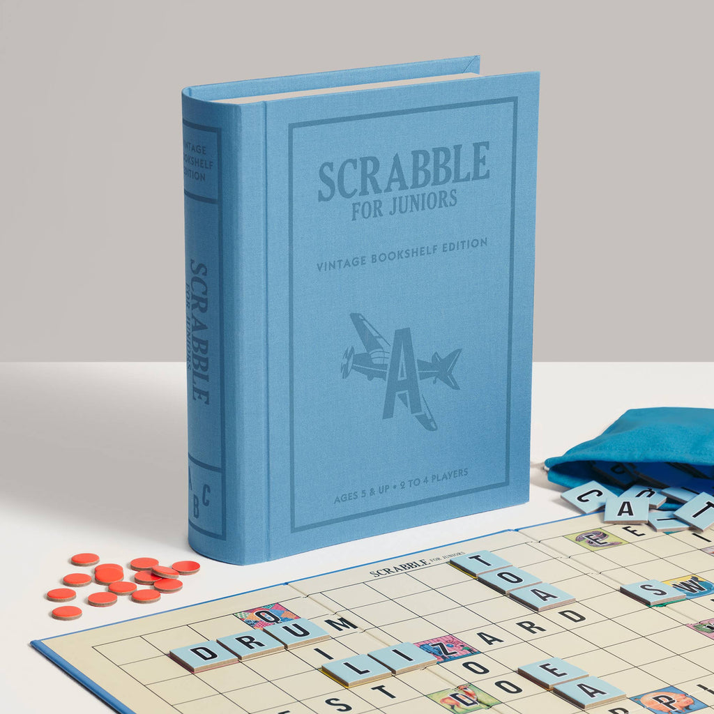 A blue Scrabble for Juniors Vintage Bookshelf Edition game box stands behind a board with letter tiles spelling “DRUM” and “LIZARD,” accompanied by red tokens and a small blue pouch.