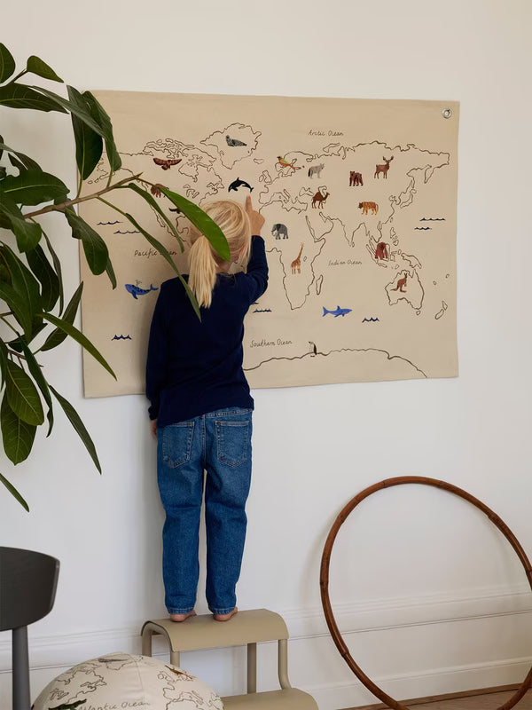 A young child stands on a small step stool, exploring the Ferm Living The World Textile Map - Off-white on the wall. A large green plant and a round wooden item nearby enhance the room’s cozy décor.