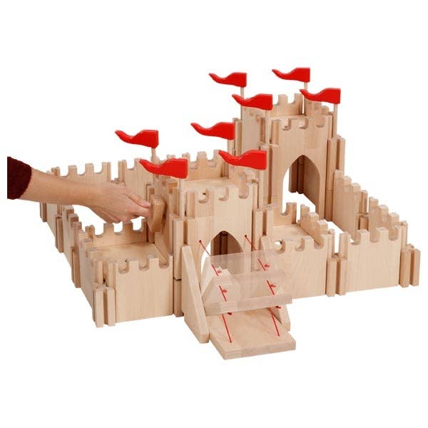 The Holztiger Knight's Castle is a handcrafted European wooden playset with red flags, a drawbridge, and detailed battlements. A hand is shown placing or adjusting a piece of the castle structure.